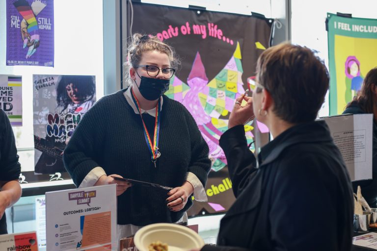a caucasian non-binary person with long mousey brown and blond hair, wearing a black mask and speaking to a community member