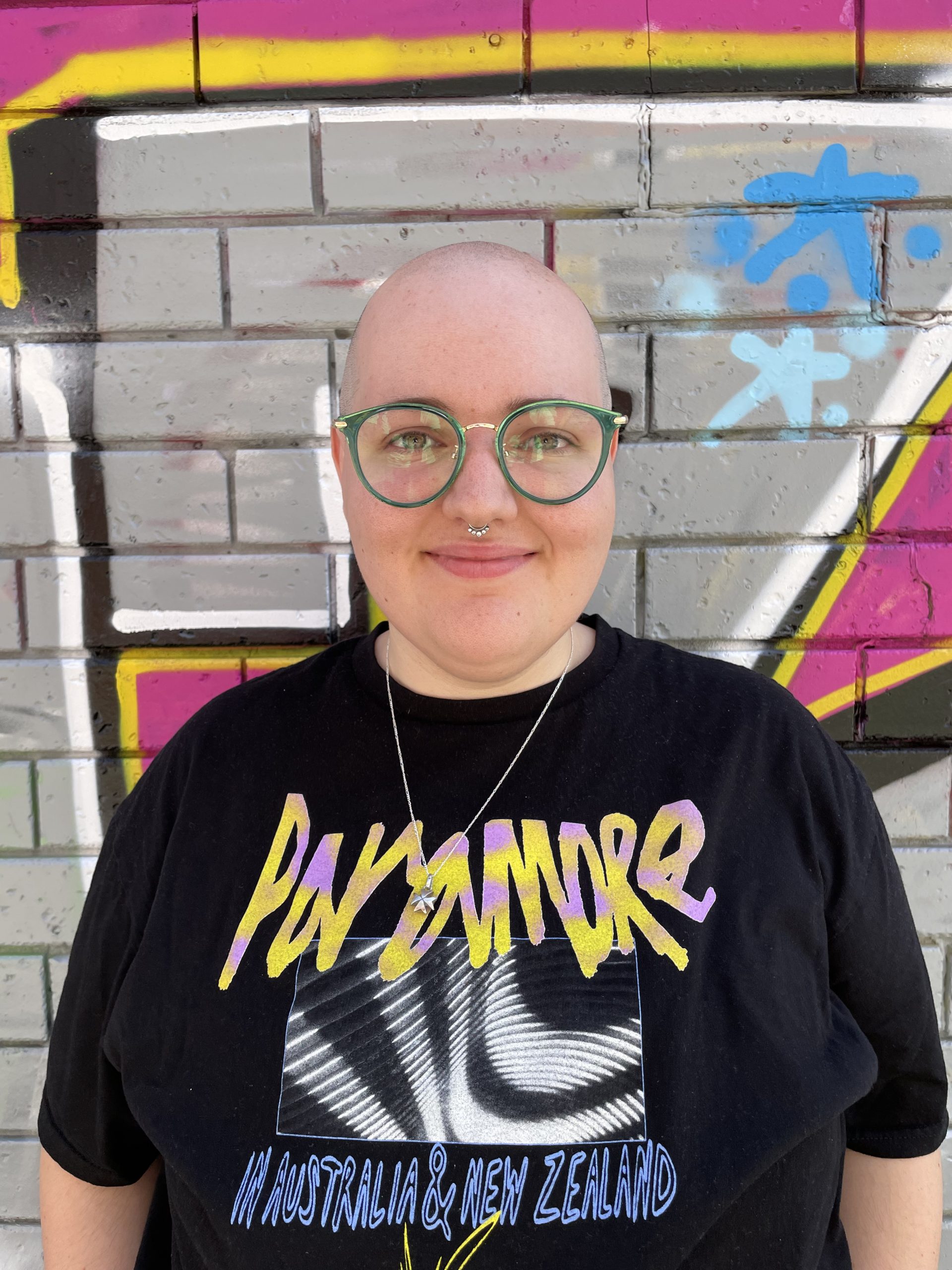 Staff member Jay standing in front of a wall of street art smiling, wearing green glasses and a band tee shirt.