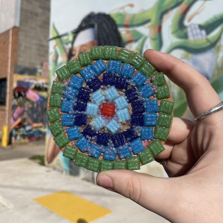 a palm-sized, round mosaic with green, blue and red tiles on a background of Jets Studios building mural (mural is an abstract side profile of a woman with long black hair)