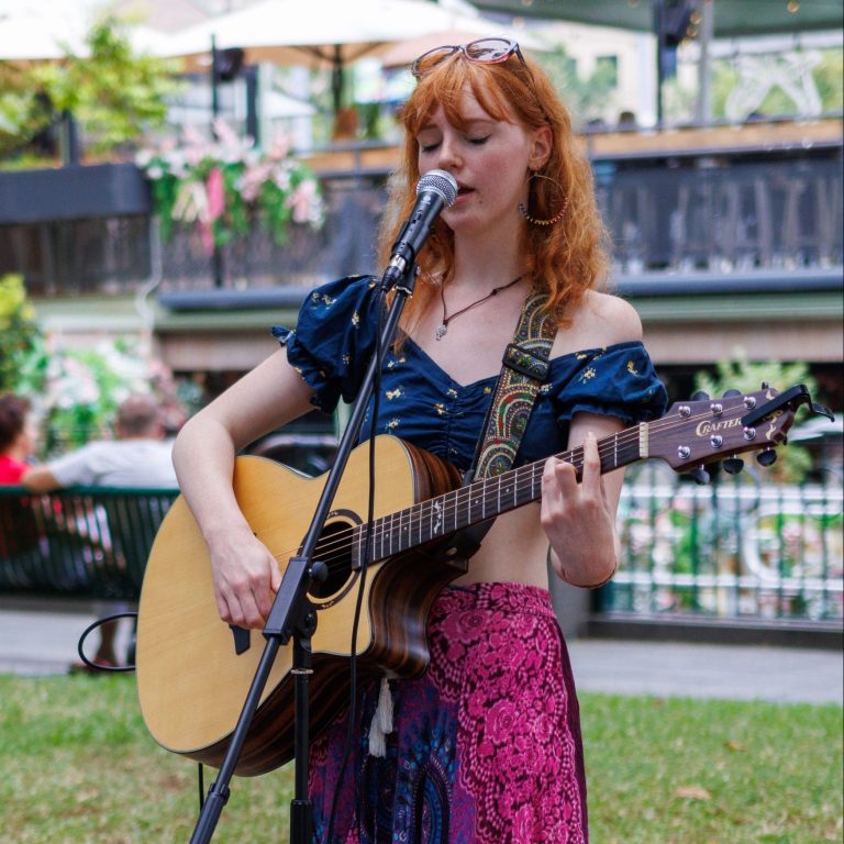 Photo of Lily playing a guitar and singing during one of her busking gigs in Melbourne. Lily wears a colourful top and skirt, and has vibrant red hair.