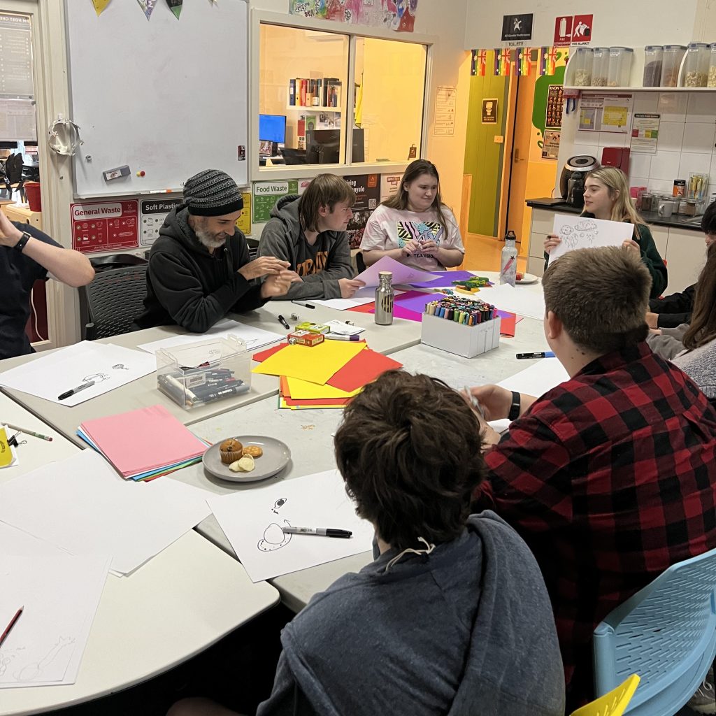 A group of young people (or various genders) sitting around an oval table, collaborating on art.