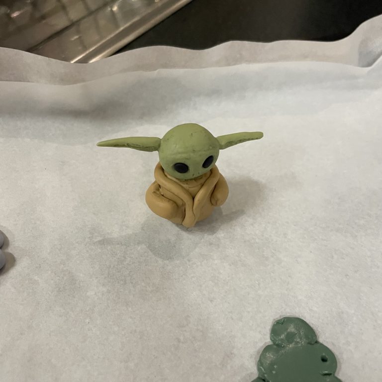 A polymer clay figure resembling character Baby Yoda. Baby Yoda is sitting on a baking sheet.