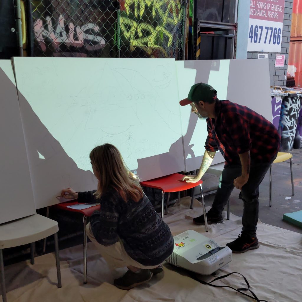 Two adults (a mand and woman) crouching or bending over to reach to sketch on large canvases. There is a projector screen projecting the outline onto the canvas, which the two people are tracing.