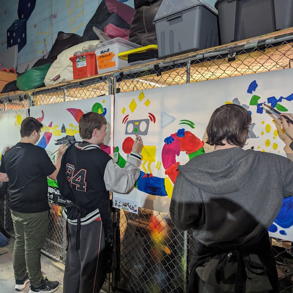 3 young adult men all working on painting large canvases. There is lots of colour on the canvases, and the prominent feature is a multi-colour waterslide.