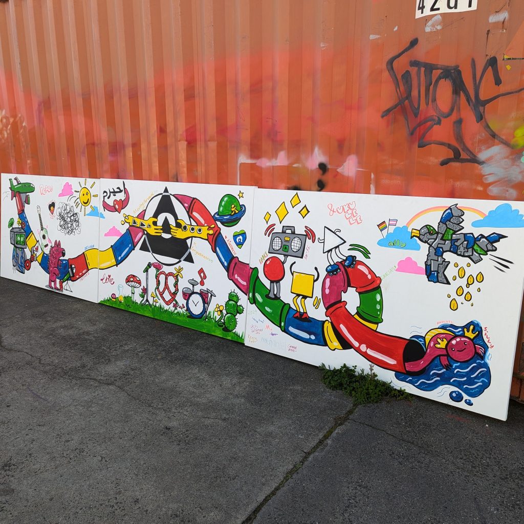 A mural made up of 3 colourful and large canvases, created with acrylic paint. The 3 large canvases are lined up against an orange shipping container, and the prominent feature is a multi-colour waterslide.